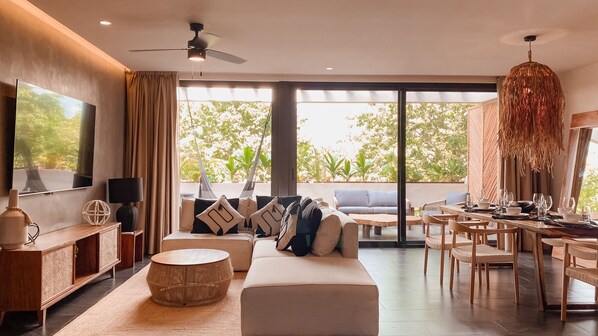 Een moderne woonkamer met een flatscreen televisie, een plafondventilator, een zithoek, een eethoek met stoelen en een tafel, en grote ramen met uitzicht op groen.