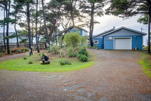 A residential property with a blue garage, a gravel driveway, and a fenced yard with trees and shrubs. - Oceanfront Home in Port Orford: The Lighthouse (Port Orford)