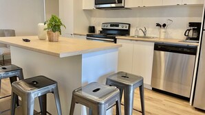 A modern kitchen with a central island, stainless steel appliances, and bar stools. - Appart-Hôtel L'UNI (Québec City)