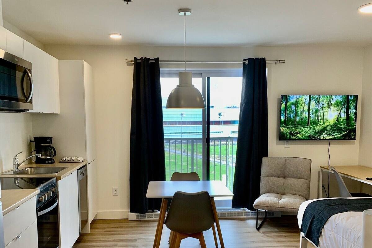 A modern kitchen with a microwave, coffee maker, and dining table. There is a TV mounted on the wall and a sofa in the living area.