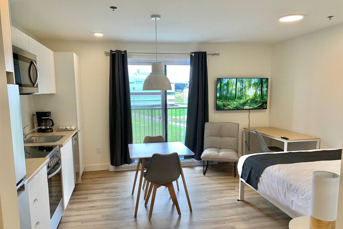 A modern hotel room with a kitchenette, a dining area, a bed, and a TV.