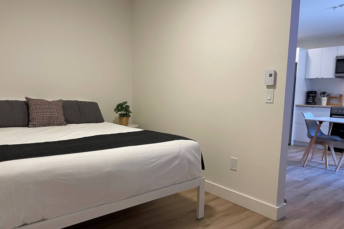 A bedroom with a bed, a small bedside table, a potted plant, and a view into a kitchen area.