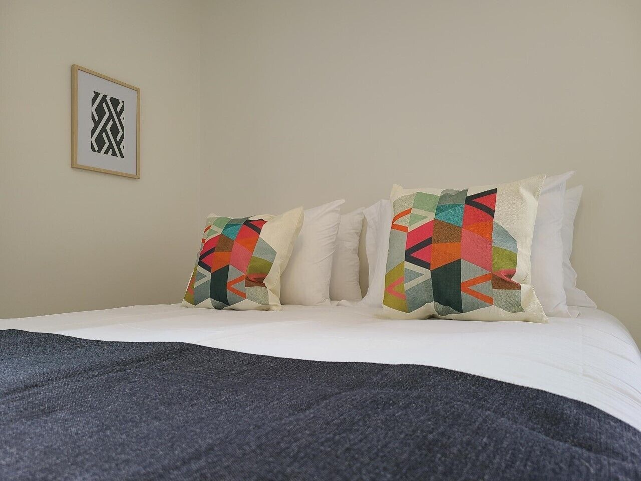 A bed with white linens and two colorful pillows, a framed abstract artwork on the wall, and a dark gray throw blanket.
