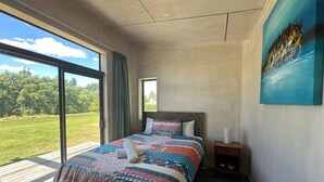A bedroom with a large bed, a nightstand, a window with a view of a green landscape, and a painting on the wall. - The River Cabin, Twizel. (Twizel)