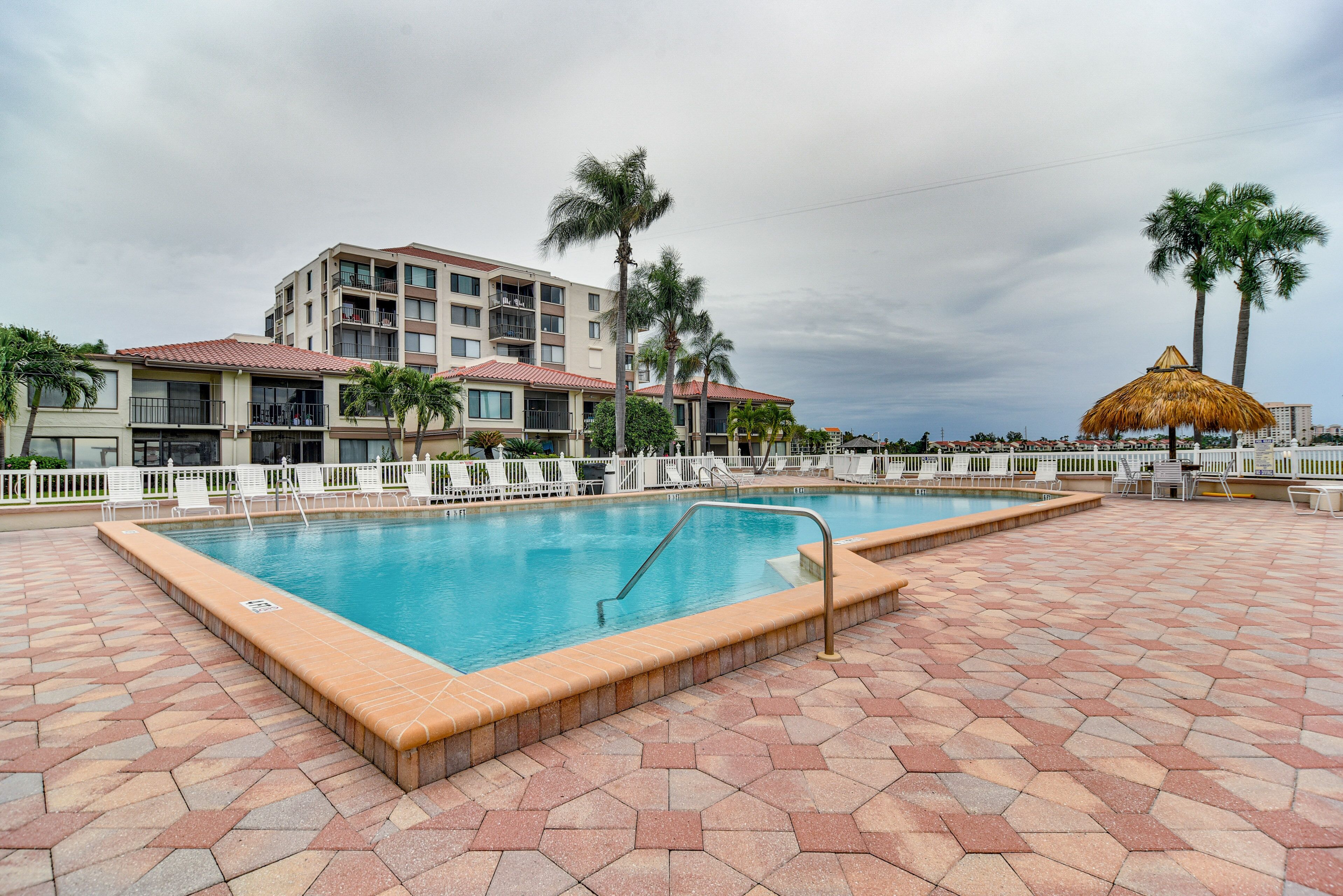 A swimming pool with a tiled deck, surrounded by palm trees and a building with balconies.