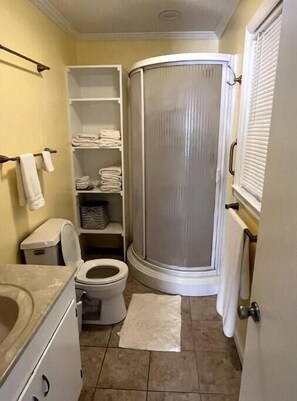 A compact bathroom with a toilet, a shower enclosure, and a storage shelf with towels. - Harry's House (Rochelle)