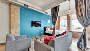 A modern hotel room with a blue accent wall, a bed with white bedding and red pillows, a TV mounted on the wall, and a city view through large windows.