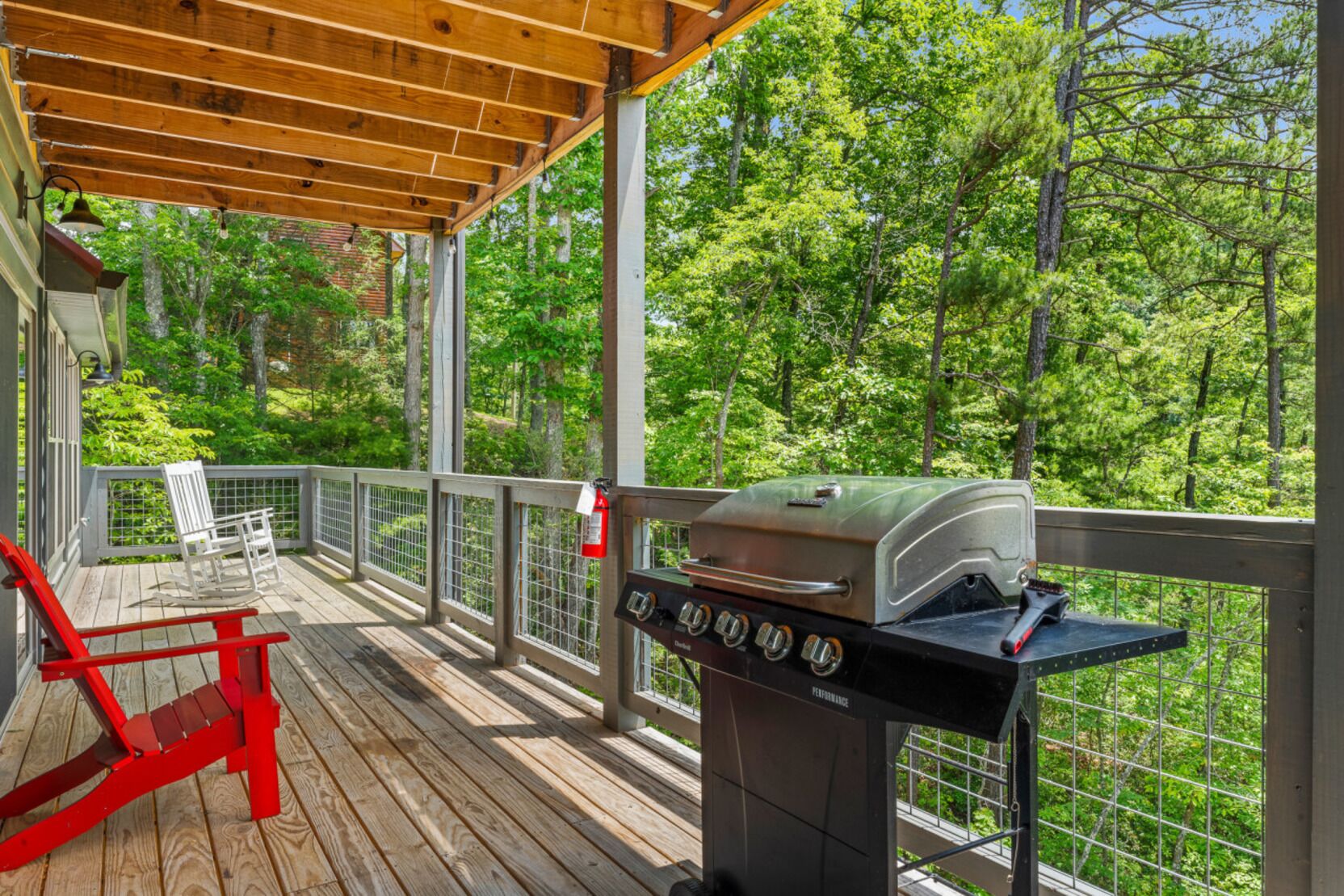 A covered outdoor deck with a barbecue grill, a red rocking chair, and a view of a forested area.