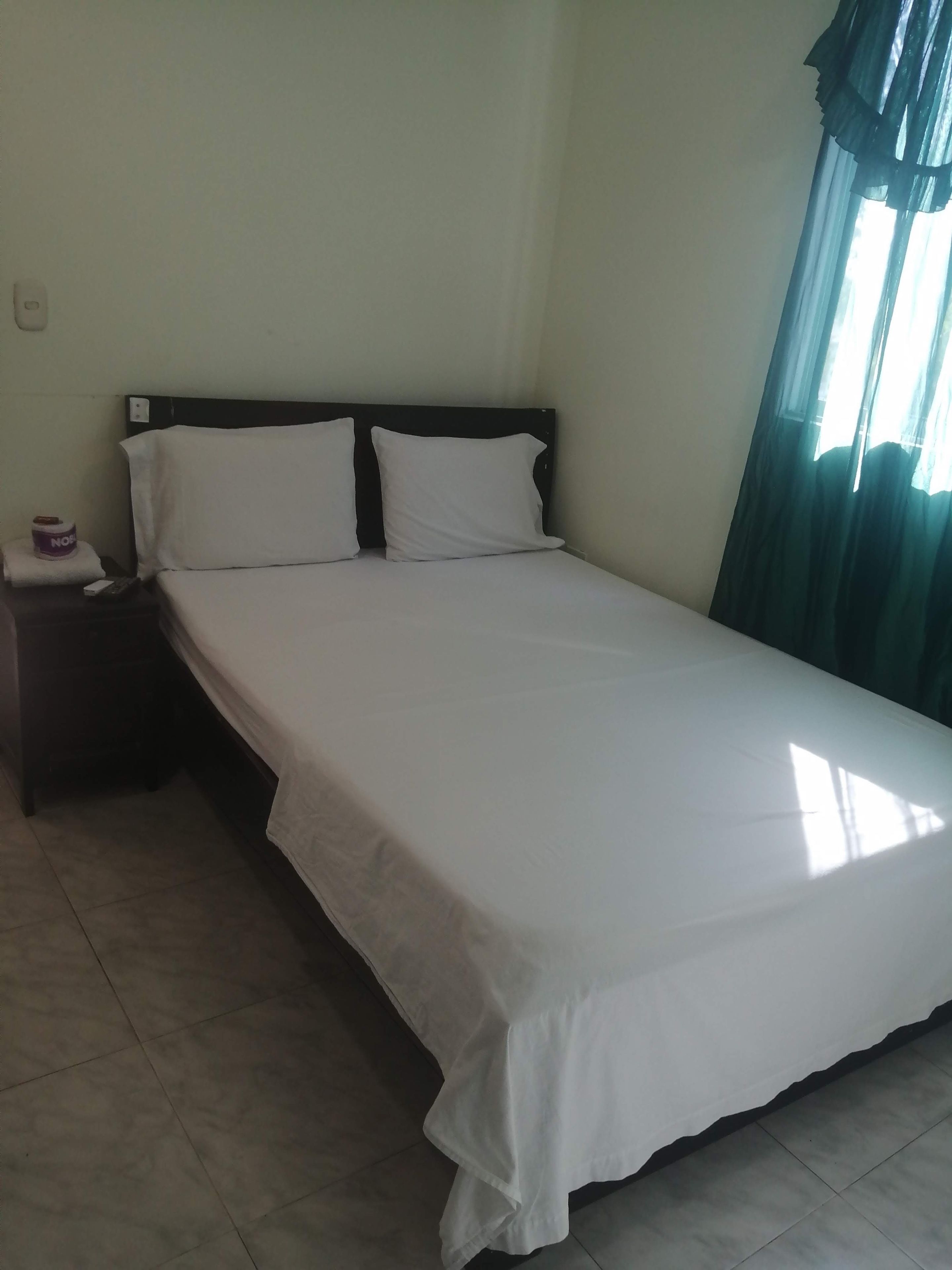 A single bed with white bedding in a room with a plain wall and a window with curtains.