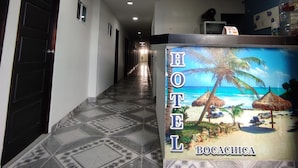 Zona de recepción de hotel con suelo de baldosas, un mostrador con un mural de playa tropical y un pasillo con puertas y ventanas.