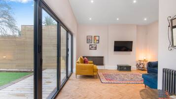 A spacious living room with a large sliding glass door leading to a patio, a yellow sofa, a blue armchair, a television, and a radiator.