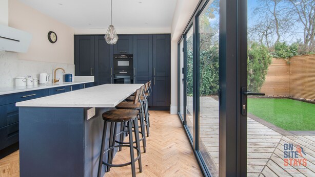 A modern kitchen with a large island, dark cabinetry, and a view of a garden through large sliding glass doors.