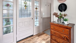 Un pasillo con piso de madera, una puerta blanca con vidrieras, un aparador con cajones y una pared decorada con sombreros y una vela.
