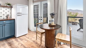 Una cocina con un refrigerador blanco, armarios azules y una mesa de comedor de madera con sillas. Hay una ventana con cortinas y vistas al exterior.