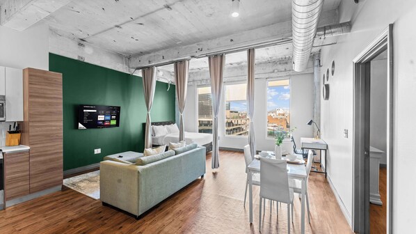 A modern apartment with a kitchenette, a sofa, a dining area, and a large window with a city view. - 3BR Loft Near Convention Center + Breakfast + Pool (Dallas)