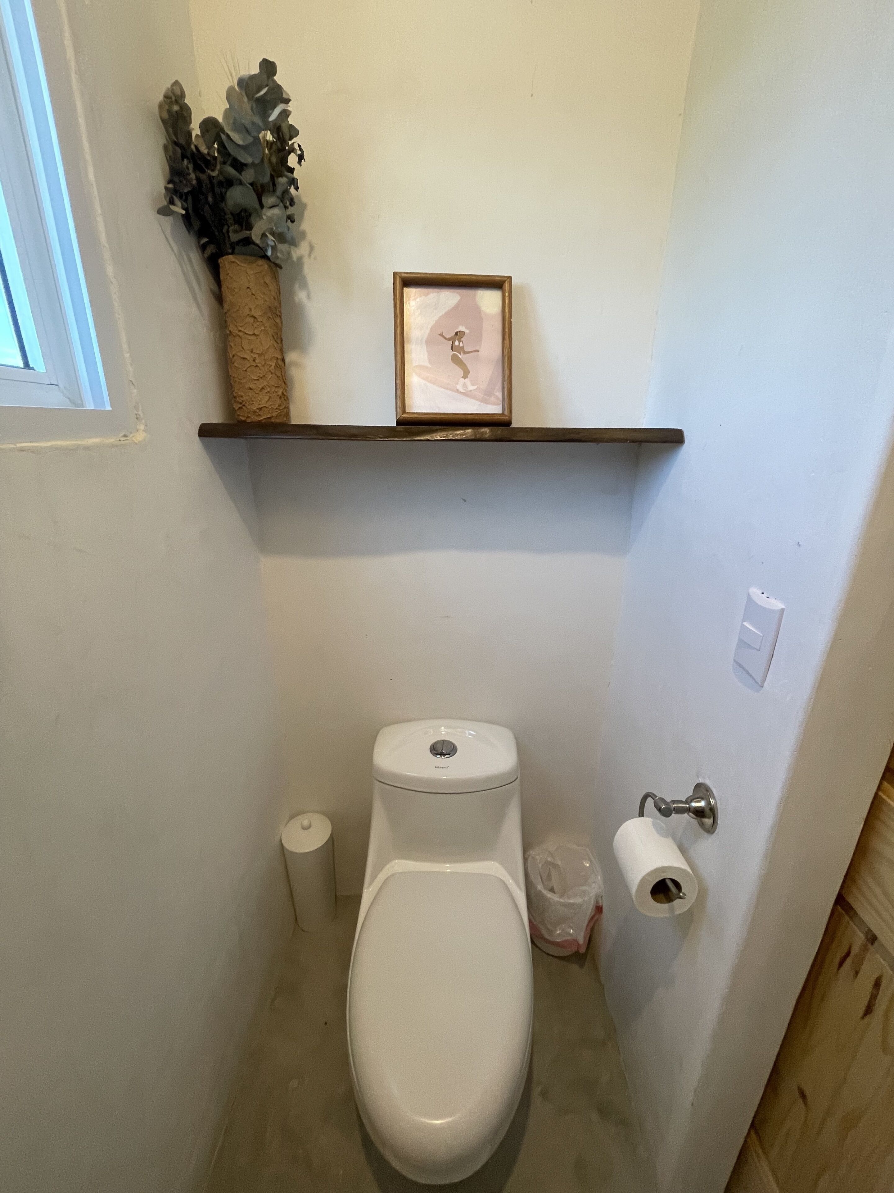 A small bathroom with a toilet, a shelf with a framed picture, and a roll of toilet paper.