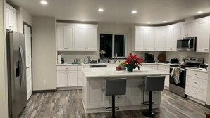 A modern kitchen with white cabinets, a large island with bar stools, a stainless steel refrigerator, and a microwave.