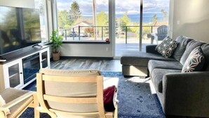 A living room with a large TV, a sofa, a chair, and a balcony with a view of the outdoors.