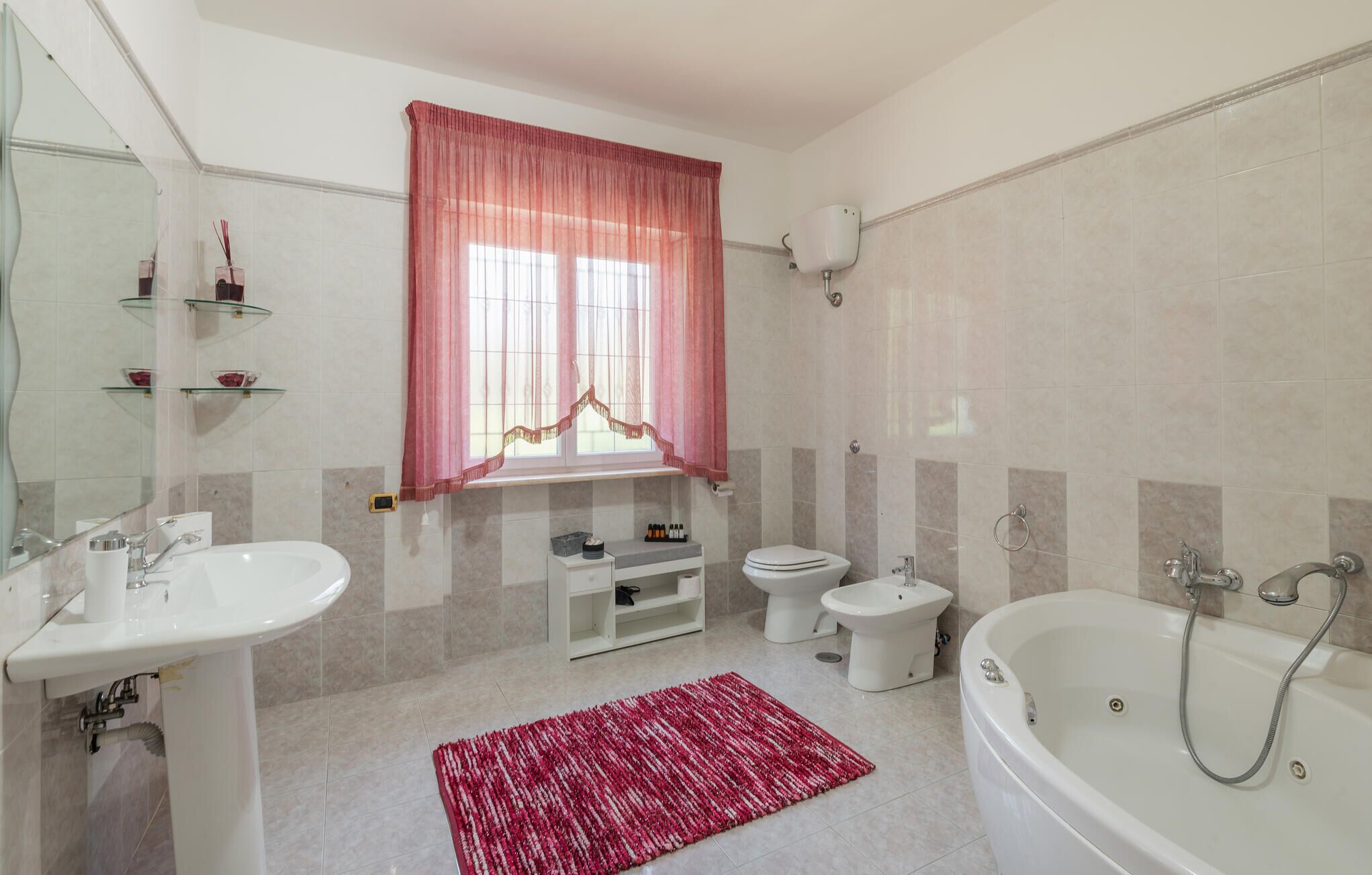 A modern bathroom with a bathtub, a toilet, a sink, and a vanity. There is a window with pink curtains, a mirror, and a red rug on the floor.