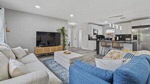 A modern living room with a sofa, ottoman, and a flat-screen TV. A kitchen with a dining area is visible in the background.