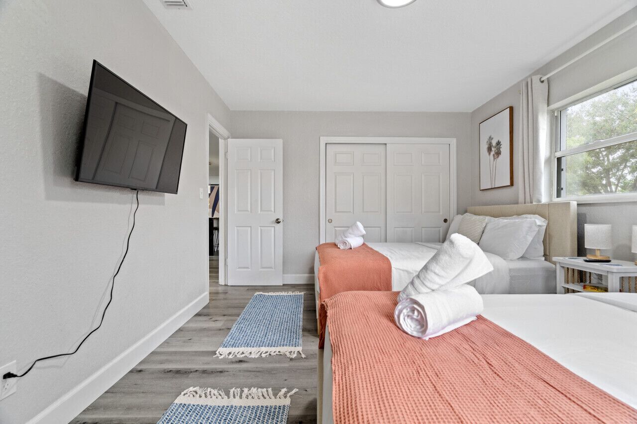 Habitación de hotel con una televisión de pantalla plana en la pared, una cama con ropa blanca y una manta rosa, una segunda cama con ropa blanca, una mesita de noche con una lámpara y una ventana con vistas a árboles.