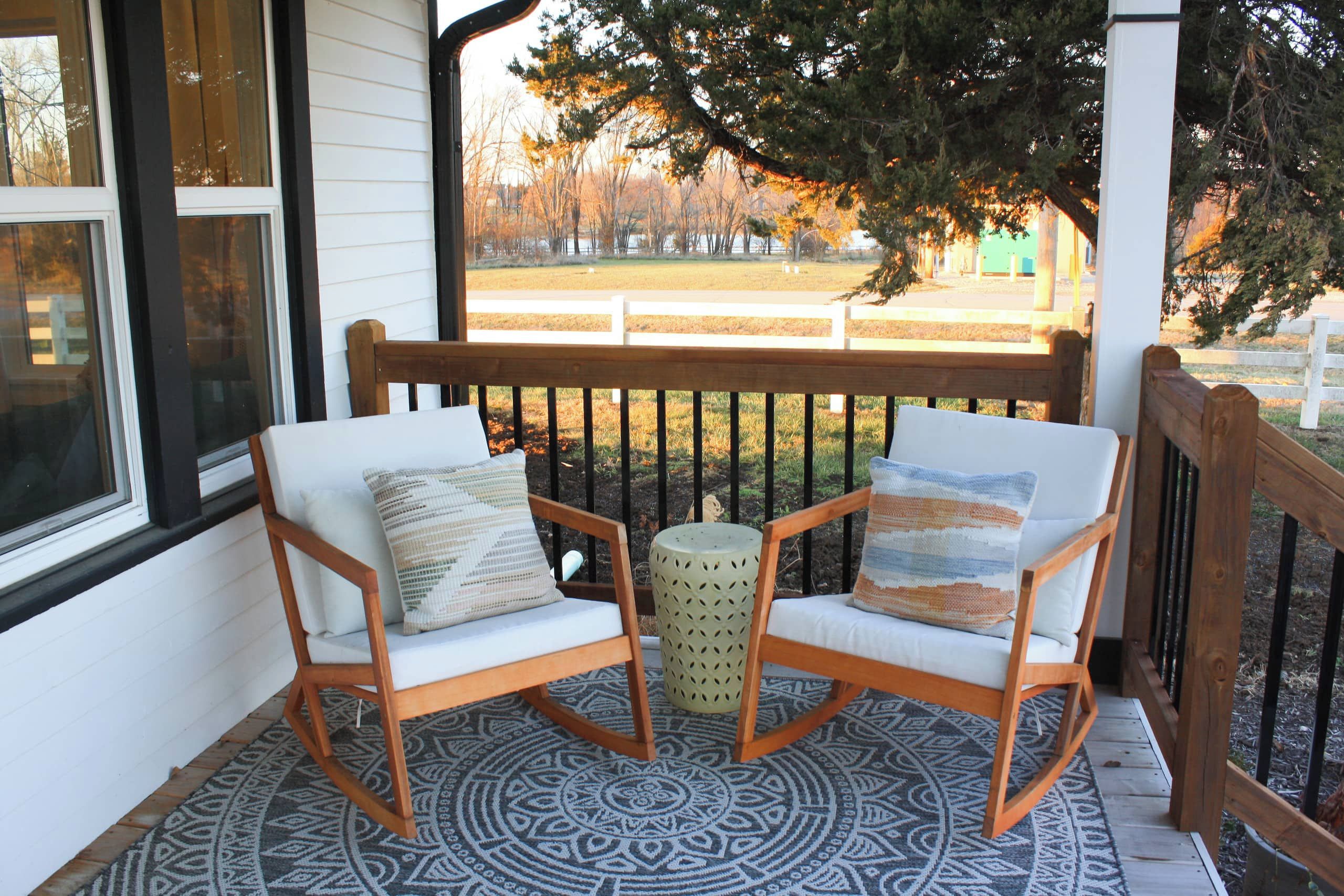 A porch with two wooden chairs and a small round table, a patterned rug, and a view of a fenced yard with trees.