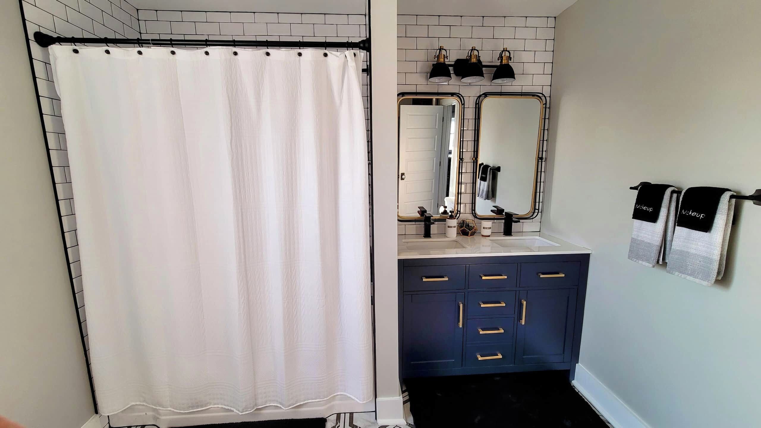 Un baño con cortina de ducha blanca, un lavamanos con espejo y un mueble azul oscuro con tiradores dorados.