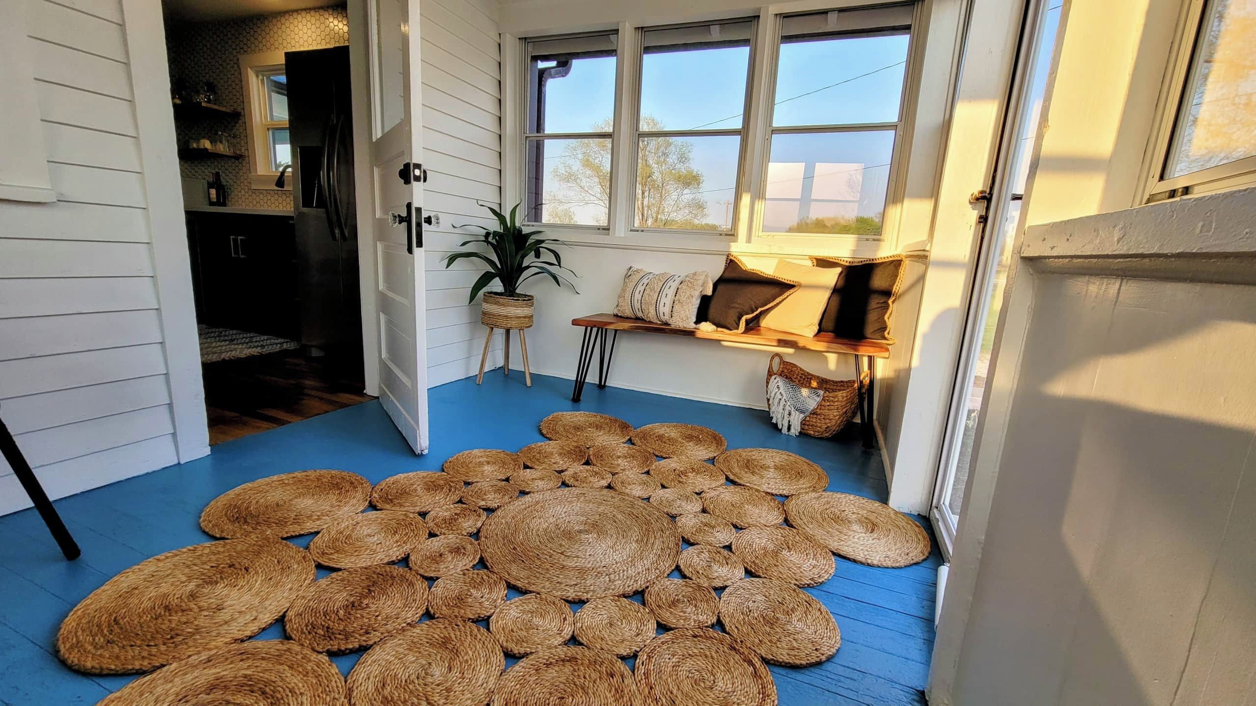 Ein Zimmer mit blauem Boden, einem großen gewebten Teppich, einer Kübelpflanze und einer Bank mit Kissen.