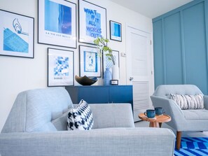 Una sala de estar moderna con un sofá gris, un sillón azul y una pequeña mesa de madera. En la pared hay obras de arte enmarcadas y un mueble azul con una planta.