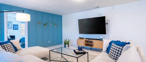 A modern living room with a large flat-screen TV, white sofas, a glass coffee table, and blue accents.