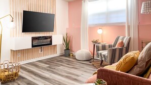 A modern living room with a flat-screen TV mounted on a wooden panel wall, a comfortable sofa, a striped armchair, and a dining table with a lamp. - [1] StayAwhile: The Pink Retreat. 2-Min Walk to Mass! (Lawrence)