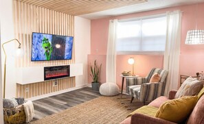 A living room with a flat-screen TV, a fireplace, a striped armchair, a round ottoman, and a small side table with a lamp. - [1] StayAwhile: The Pink Retreat. 2-Min Walk to Mass! (Lawrence)