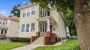 A two-story residential building with a front porch, a red door, and a brick foundation.