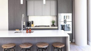 Une cuisine moderne avec des meubles de couleur grise, un plan de travail blanc et une cuisinière encastrée. On trouve quatre tabourets en bois près du bar.