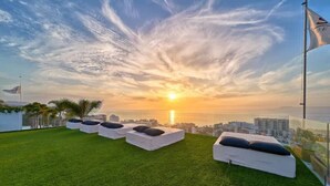 A rooftop terrace with white cushioned seating and a view of the sunset over the ocean.