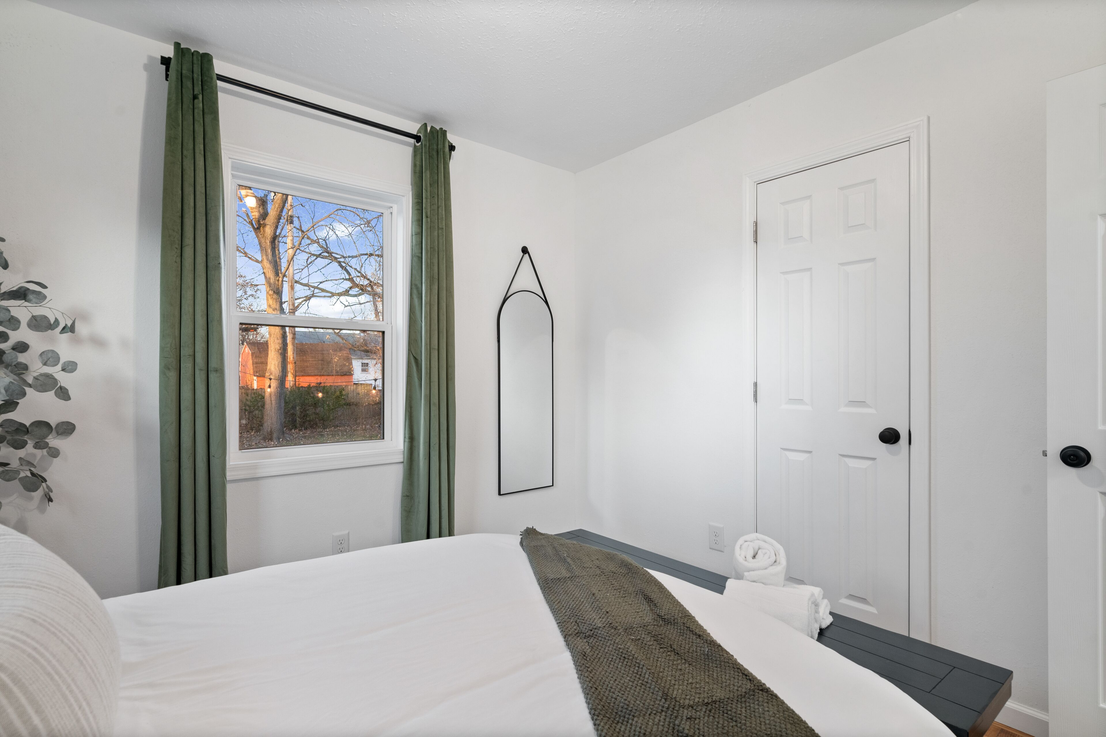A bedroom with a bed, a window with curtains, a door, and a wall-mounted mirror.
