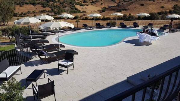 A pool area with lounge chairs and umbrellas, surrounded by a fence and hills in the background.