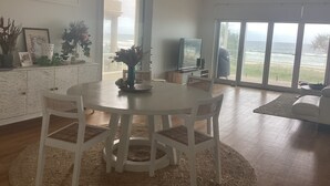 Uma sala de jantar com uma mesa redonda e cadeiras, uma TV e vista para o oceano através de grandes janelas.