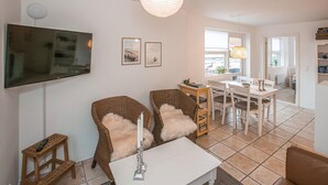 A living room with a TV, wicker chairs, a dining table, and a white table lamp.