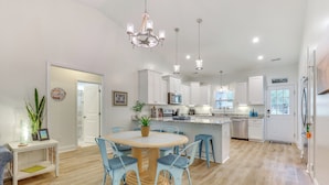 A modern kitchen with white cabinets, a central island, and a dining area with a round table and blue chairs. - Serene Oak Island Home w/ Private Saltwater Pool! (Oak Island)