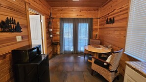A wooden cabin interior with a kitchen area, a dining table with chairs, and a small stool.