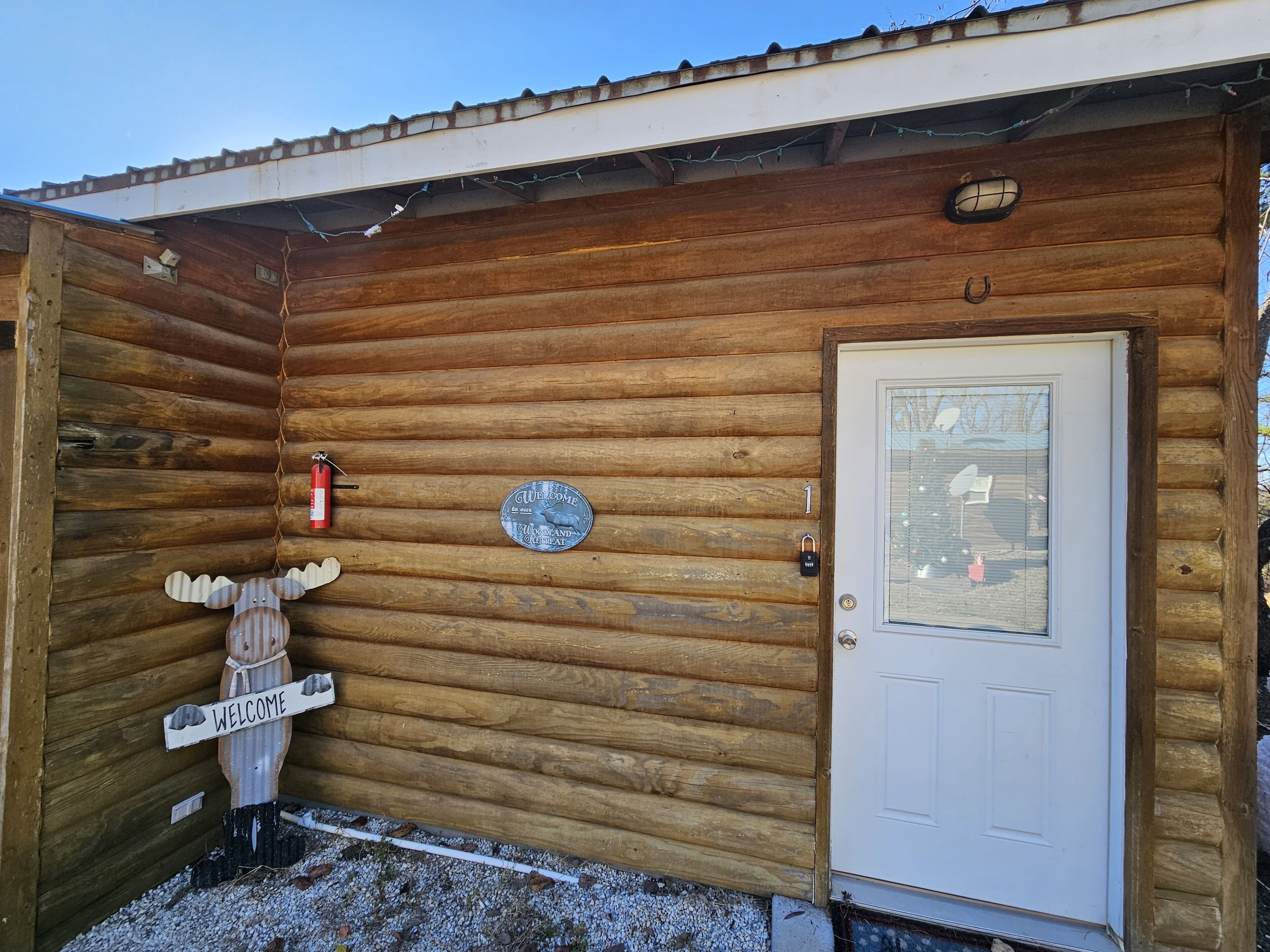 A wooden cabin with a white door, a welcome sign, a fire extinguisher, and a number plate.