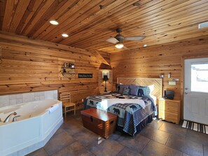 Une chambre dans un chalet en rondins, avec un grand lit, une tête de lit en bois, une table de chevet, une commode, un banc en bois, une grande fenêtre et un ventilateur de plafond.
