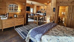 A cozy wooden cabin interior with a large bed, a dining area, a kitchen, and a bathroom.