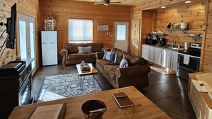 A cozy cabin interior with a wooden ceiling, a living area with a sofa and coffee table, a kitchen with appliances, and a dining area with a table and chairs.