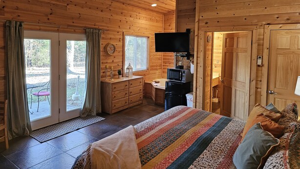 A wooden cabin bedroom with a bed, dresser, television, and a view through the door. - Cabin-style private suite: king bed, jetted tub, kitchenette, Wi-Fi & private patio—near Beaver Lake & Eureka Springs. (Eureka Springs)