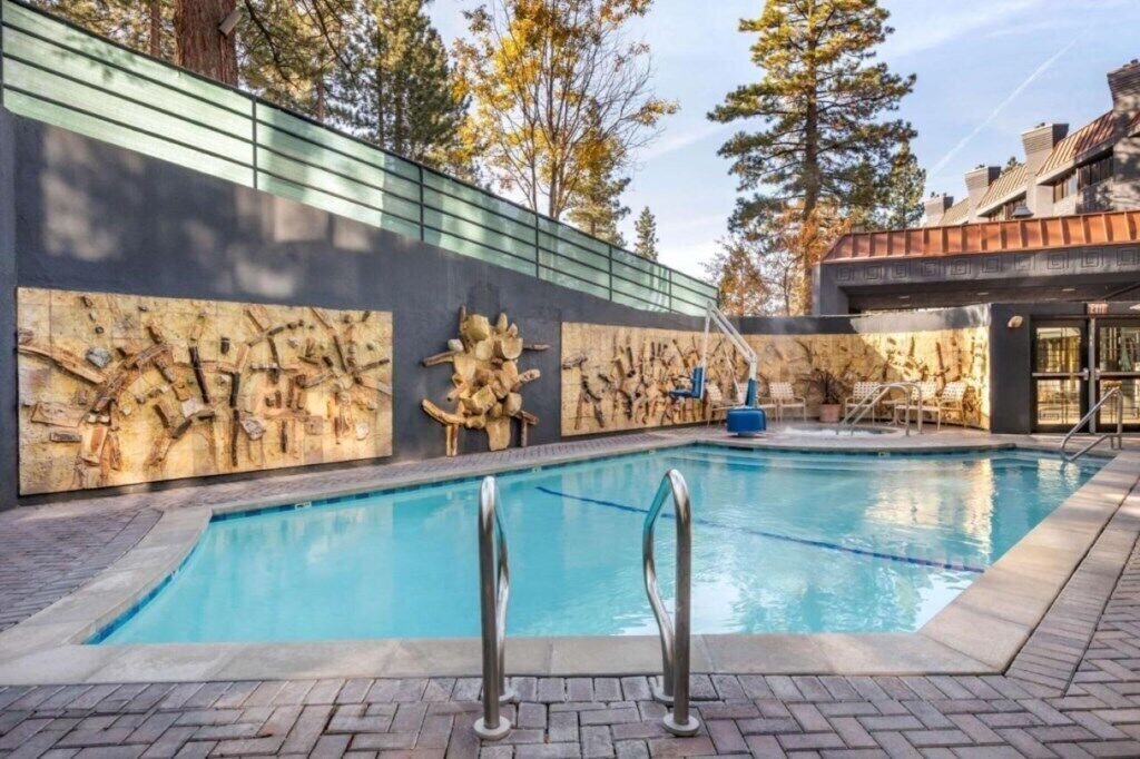 Une piscine ornée d’une fresque murale en pierre, un espace de détente avec des fauteuils et un bâtiment en arrière-plan.