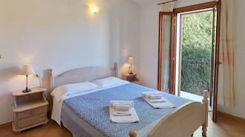 A bedroom with a bed, two bedside tables with lamps, a window with curtains, and a view of greenery outside.
