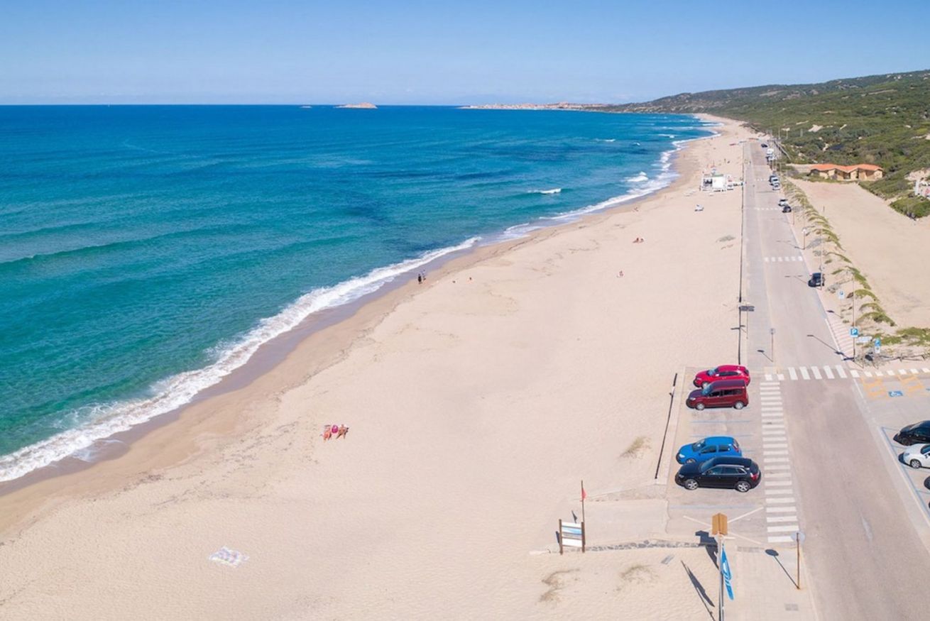 A beach with a road running parallel to it, lined with parked cars and a few people walking.