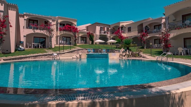 Una piscina rodeada de edificios de varios pisos con balcones, cielo despejado y algo de vegetación.